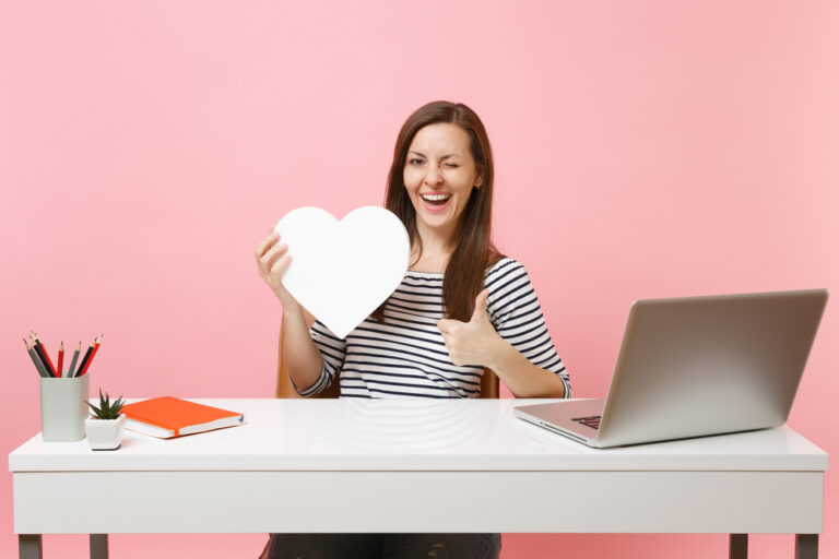 excellent customer service - Funny woman blinking showing thumb up holding white heart with copy space working on project while sit at office with laptop isolated on pink background.