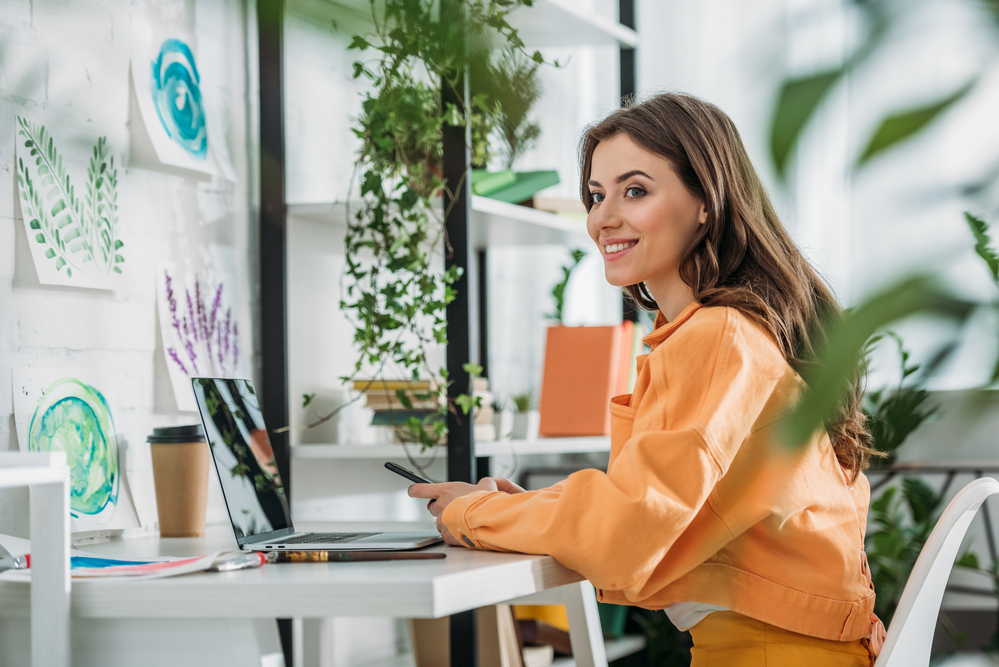 Business Decisions for Improved Well-being office - young woman in soft orange top at home office desk with computer, green plants and a smile of success and contentment