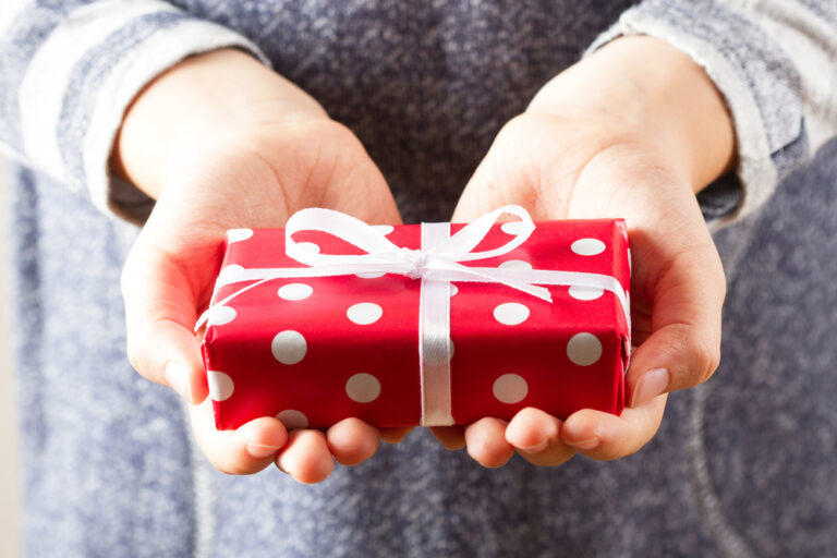 Woman in gray sweater holding gift out in hands representing the gift of Creative Strategies for Supporting Your Well-Being